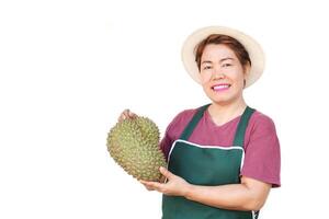 Happy Asian woman wears hat, green apron, holds durian fruit. White background. Concept, fruit seller, gardener occupation. Durians,seasonal fruits and popular. photo