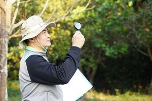 Asian man ecologist is exploring ecological system in forest, holds magnifying glass and paper notepad. Concept, examine, explore, research, study about environment, plants and nature. photo