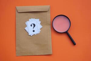 Brown envelope,magnifying glass and paper cards of question marks,orange background. Concept. Teaching aid. Education materials for doing activity or playing investigation games about find answers. photo