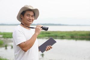 hermoso asiático hombre ecologista es topografia naturaleza a el lago, sostiene prismáticos y inteligente concepto de tableta, naturaleza exploración. ecología estudiar. pasatiempo actividad, estilo de vida. hombre explorar ambiente foto