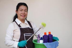 Asian woman housewife wears gloves, apron, holds bucket, bottles of cleaning liquid, alcohol spray, sanitary products and equipment for cleaning. Concept, Cleaning time for hygiene. Housekeeping. photo