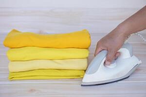 Stack of folded yellow clothes and hand holds electric iron on table. Concept, daily chore, household. ironing and folded clothes for neat and clean. Maintenance and keep garments. photo