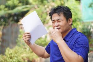 Asian man feel hot, unwell, use menthol inhaler to smell, hold paper to fan for relief symptom from hot weather. Concept, self first aid to protect from heat stroke. Refreshing. photo