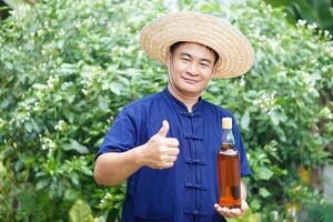 Asian man farmer wears hat, blue shirt and holds bottle of honey at his garden. Concept, Organic agriculture product from bee raising farm or nature sources. Thai local lifestyle. Natural beverage. photo