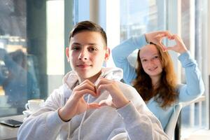 chico Adolescente niña sentar en un café por el ventana hacer manos corazón gastar hora juntos celebrar un día apagado formación ninguna publicidad gracioso y agradable real niños personas adolescencia foto