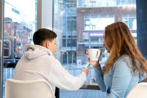 Fashion couple of young lovers at cafeteria Handsome man whisper tender kiss to pretty woman Relationship boyfriend and girlfriend together teenagers on first date near glass window coffee and phone photo