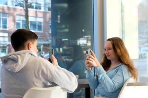 life of teenagers on the phone a boy and girl sit at table by window each with their smartphone spend time on Internet in social networks cafe School cafeteria take pictures of each other shoot s photo