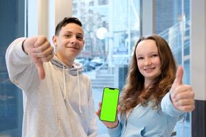 emotions boy holding thumbs down girl holding thumbs up Like it or dislike Green chroma key background on phone screen near window teenagers approve and disapprove something What can be written photo