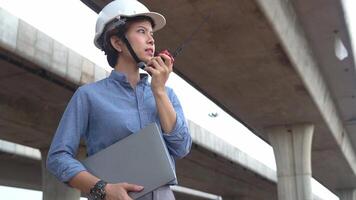 asiatico femmina ingegnere utilizzando walkie talkie a costruzione luogo in piedi vicino il calcestruzzo strada, autostrada ponte. video