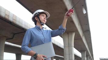 asiatico femmina ingegnere utilizzando walkie talkie a costruzione luogo in piedi vicino il calcestruzzo strada, autostrada ponte. video