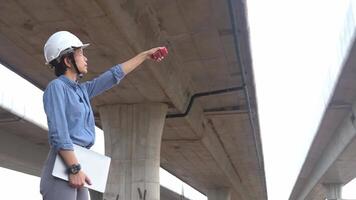 asiatico femmina ingegnere utilizzando walkie talkie a costruzione luogo in piedi vicino il calcestruzzo strada, autostrada ponte. video