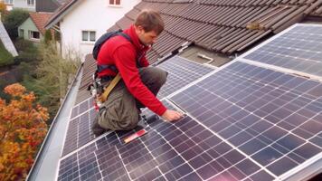 une bricoleur est installation solaire panneaux sur le sien le genou sur le toit de une allemand maison portant une rouge costume. video