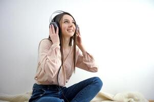 a girl in blue jeans sits on the floor on blanket on the bed raised her head up listens to music on headphones place for text online work conference attractive girl with headphones on gray background. photo