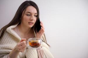sick elegant 20 years old housewife in roll neck sweater and cardigan drinking cup of hot tea with ginger, lemon and honey against winter light blue background. photo