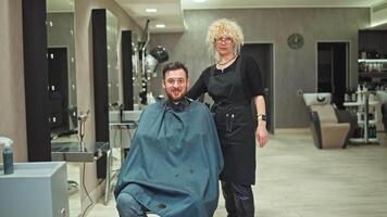 retrato do uma fêmea cabeleireiro e uma feliz homem depois de uma corte de cabelo dentro uma beleza salão. a trabalhos do uma profissional é para cortar cabelo, levar Cuidado do a aparência do beleza. Alto qualidade 4k cenas video