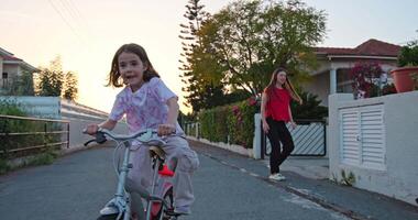 A smiling baby girl rides a bicycle on the street under the supervision of her mother. Happy childhood, active lifestyle of happiness. High quality 4k footage video