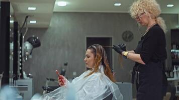 een vrouw is sms'en Aan de telefoon terwijl hebben haar haar- geverfd in een schoonheid salon. levensstijl werk van een kapper met haar- verven. hoog kwaliteit 4k beeldmateriaal video