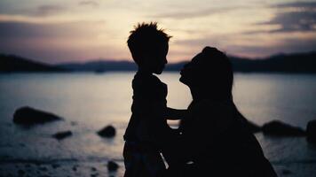 mãe Beijos a paredes às pôr do sol perto a mar. silhueta do uma criança e uma mulher, uma feliz família juntos dentro uma lindo panorama video