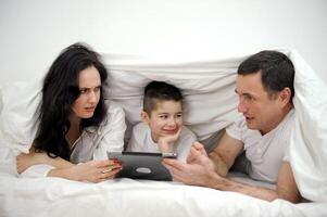 people, family and technology concept mother, father and little boy with tablet pc computer in bed at home or hotel room. Happy family spend time together lying under white blanket on bed at bedroom photo