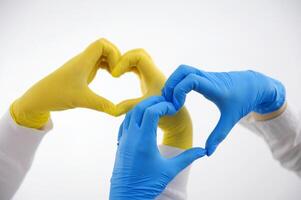 el manos de dos enfermeras hacer corazón en el aire en blanco antecedentes azul y amarillo látex guantes comprensión ayuda mutuo asistencia hipocrático juramento ayuda paciente amor de vida cardiología proteger corazón foto