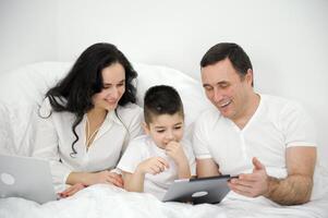people, family and technology concept mother, father and little boy with tablet pc computer in bed at home or hotel room. Happy family spend time together lying under white blanket on bed at bedroom photo