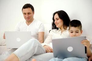 último tecnología artilugio mamá y hijo jugando en tableta padre sentado en blanco cama trabajando en ordenador portátil remoto trabajo persona de libre dedicación vacaciones familia combinar trabajo y gastar hora con familia juntos foto