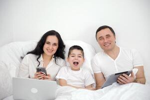 contento familia y digital tecnología. familia padres y hijo acostado en el cama utilizando inteligente Los telefonos éxito victorioso. foto