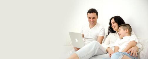 banner family husband wife and child sitting watching movie on laptop on bed relaxing together white clothes chatting online with relatives mom dad and son laughing smiling rejoicing happy life photo