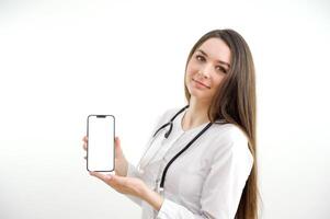 Beautiful female doctor smiling and showing a blank smart phone screen isolated on a white background. Portrait of happy female doctor promoting smart phone. photo