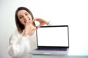 girl on a white background points to a laptop screen a girl in white shows a heart with her hands smiling near a laptop with an empty space for advertising text of any content photo