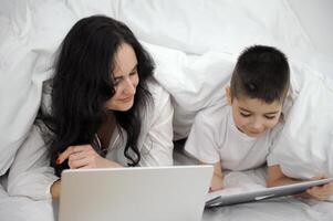 work at home online woman under blanket working on laptop beside son playing games on tablet sick at home get well family isolation quarantine white sheet duvet black hair mom and son photo