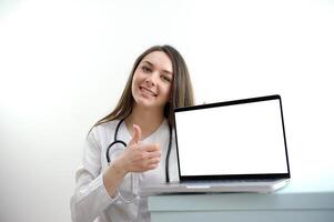 young girl doctor nurse sits near a laptop and shows thumb up ad place text advertising food supplements proper nutrition healthy way of life smile tenderness femininity white studio background photo