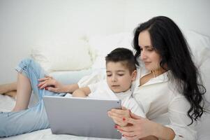 Mother and son laying down on bed and watching cartoons tablet. Bed time concept. Bedroom interior. photo