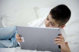 Adorable blond toddler boy laying on the sofa and playing with tablet pc at home, indoors. Child with tablet computer. photo