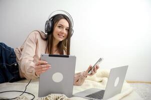 phone tablet laptop a lot of different technologies girl listens to music headphones plays phone works on computer on floor bed rest at home Work online on Internet social networks freelancer photo