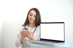 joven hembra médico mirando dentro el teléfono siguiente a un ordenador portátil con un blanco pantalla vacío espacio publicidad médico tecnología red moderno digital cirujano, enfermero, farmacia con estetoscopio foto