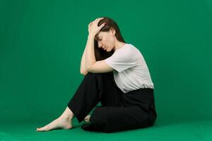 a young woman sits on a green background, holds her head with her hands, suffers, experiences, barefoot, black pants, pink T-shirt photo