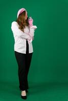 full-length woman in black pants, shoes and a pink shirt, winter gloves and a beret on her head on a green background, standing in thought, crossing her fingers on her left hand, long hair photo