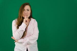 Portrait of flirty young woman making silence gesture over green background. Caucasian lady wearing shirt posing with finger on lips. Silence and secret concept photo