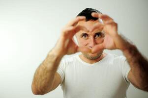 sin afeitar adulto hermoso hombre vistiendo casual camiseta en pie terminado aislado blanco antecedentes sonriente en amor haciendo corazón símbolo forma con manos. romántico concepto negro pelo con gris foto