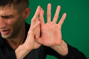 Frightened adult guy shows a stop gesture, trying to protect himself, looks to the side, closed his eyes and raises hands up, green studio background, copy space. human reactions and negative emotions photo