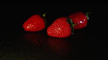 Three fresh strawberries lie on a wet surface in the dark. Black background video
