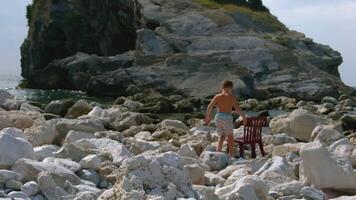 Garoto senta em cadeira de mar. criativo. Garoto anda em sozinho em rochoso costa com pedras. Garoto senta em cadeira entre pedras em costa em ensolarado dia video