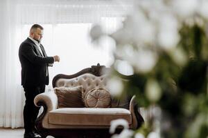 A man in a suit is standing in front of a couch, looking at his watch photo