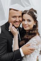 a handsome and stylish groom hugs and kisses his bride in a white dress under her veil photo