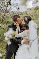 The groom kisses his bride in a white dress against the background of blooming trees in a green garden in spring photo