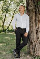A man in a white shirt and black pants stands next to a tree photo