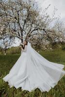 a beautiful bride in a white dress with a long veil stands near a blooming tree and holds a bouquet of flowers in her hands. Spring wedding photo