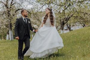 the groom and his bride are walking on the green grass in the spring garden. The bride is in a chic white dress, the groom is in a black suit photo