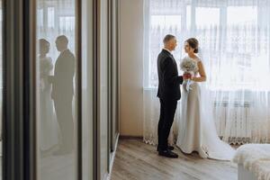 A bride and groom stand in front of a window, with the bride holding a bouquet photo
